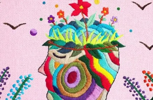 Colorful embroidered artwork depicting a stylized human head with a floral crown, surrounded by birds and vibrant plants against a pink background.