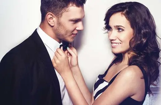 A couple in formal attire gazes into each other's eyes, with the woman adjusting the man's bow tie in an intimate, playful moment.