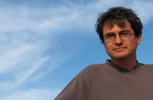 A man with dark, tousled hair and glasses stands outdoors against a bright blue sky with scattered clouds, wearing a gray shirt.