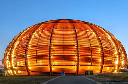 A striking spherical building with wooden slats illuminated at dusk, set against a blue sky, surrounded by greenery and flags.