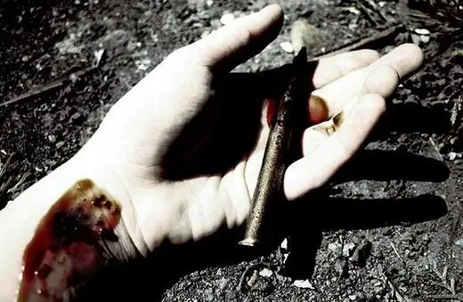 A close-up of a bloodied hand resting on the ground, holding a rusty nail, with visible injuries on the wrist and dirt surrounding.
