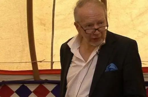 A man wearing a partially unbuttoned suit jacket speaks in a tent, with colorful patterns in the background.