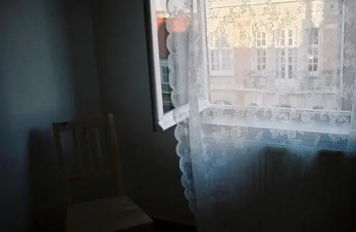 A softly lit room with a lace curtain covering an open window, revealing a glimpse of the outside buildings. A wooden chair stands nearby.