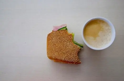 A partially eaten sandwich with sesame seeds on the bun, accompanied by a small cup of light-colored liquid, possibly a beverage.