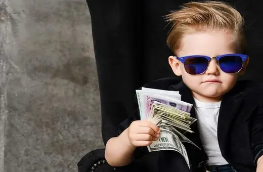 A young child wearing sunglasses and holding a stack of cash, looking confidently at the camera while seated on a chair against a dark background.
