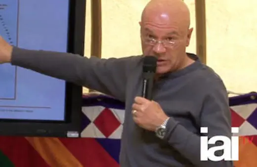 A bald man in a gray sweater gestures while speaking into a microphone, presenting information displayed on a screen in a colorful setting.