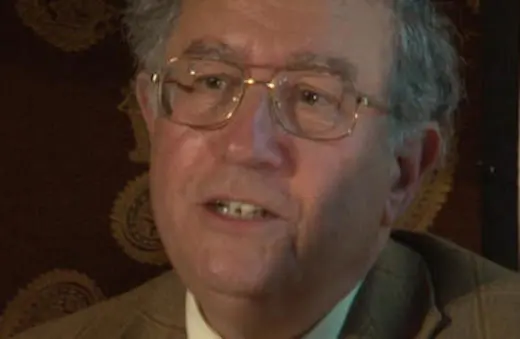 A middle-aged man with glasses is speaking, dressed in a suit, against a patterned background.