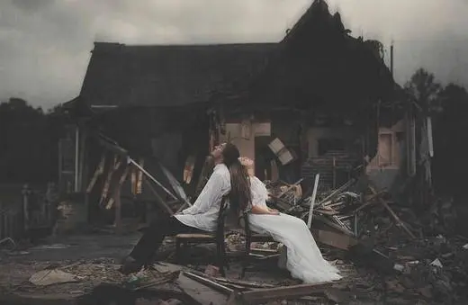 Two figures in elegant attire sit back-to-back on a chair amidst the ruins of a destroyed building, set against a moody, overcast sky.