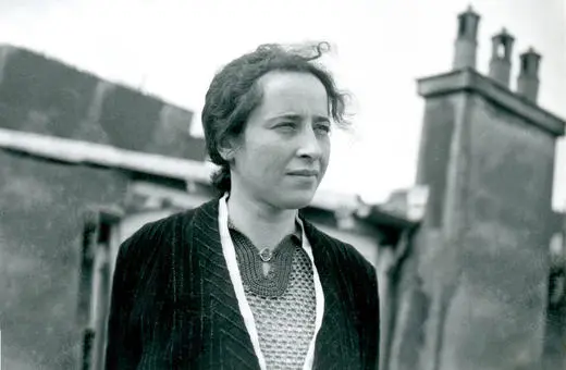 A woman stands outdoors, gazing thoughtfully, with chimneys visible in the background, captured in black and white.