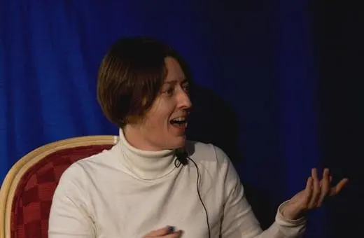 A woman with short hair, wearing a white turtleneck, is animatedly speaking while seated in front of a blue backdrop.