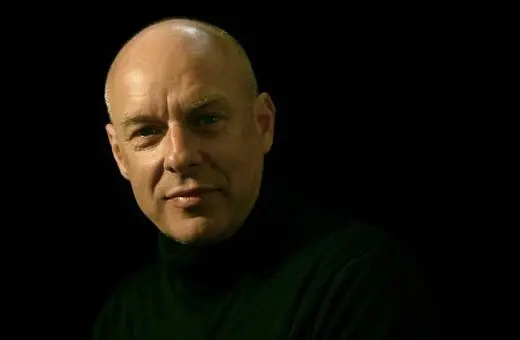 A bald man in a black turtleneck smiles subtly against a dark background, exuding a contemplative and artistic presence.