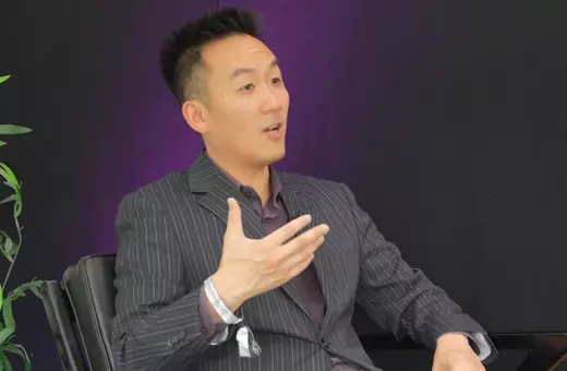 A man in a pinstriped suit gestures while speaking in a seated position, with a dark background and a plant to the left.
