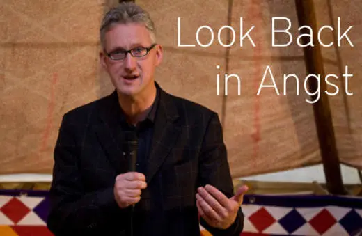 A speaker with glasses gestures while holding a microphone in front of a colorful backdrop, promoting an event titled 