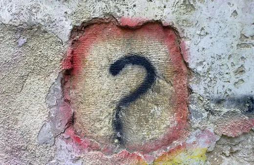 A large, black question mark is painted on a textured, weathered wall with patches of red and beige around it.