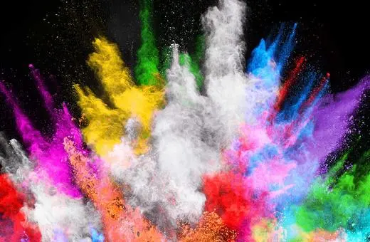 A vibrant explosion of colored powders in red, yellow, blue, green, purple, and white against a black background, creating a dynamic and energetic visual effect.