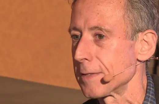 A man with short gray hair speaks at a podium, wearing a microphone headset, with a focused expression.