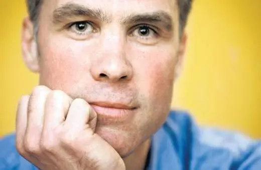 A close-up portrait of a thoughtful man resting his chin on his hand, with a neutral expression against a bright yellow background.