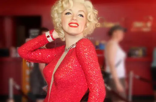 A glamorous figure resembling a classic Hollywood star, dressed in a sparkling red gown with a playful pose, set against a vibrant red background.