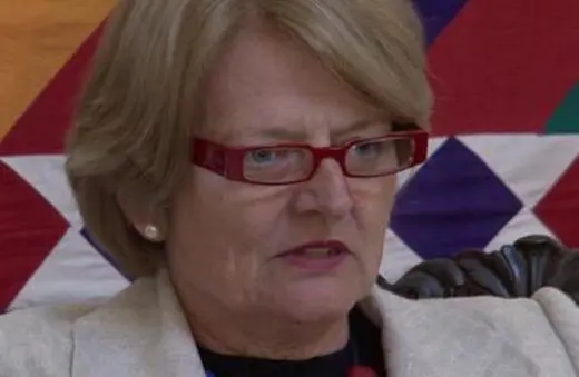 Close-up of a middle-aged woman with short blonde hair and red glasses, speaking intently while seated against a colorful geometric background.