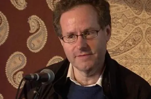 A man with glasses speaks into a microphone, against a patterned background. He appears engaged in a discussion or presentation.