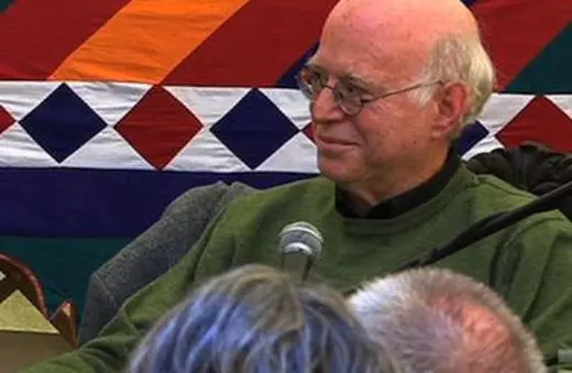 A man with glasses and a bald head sits comfortably, wearing a green sweater, in front of a colorful patterned backdrop, engaged in conversation.