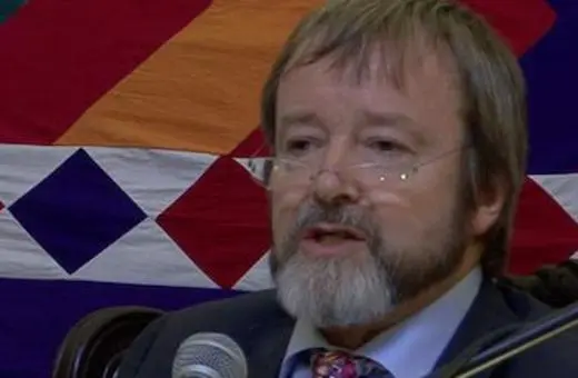 A man with a beard and glasses speaks into a microphone, seated against a colorful patterned backdrop.