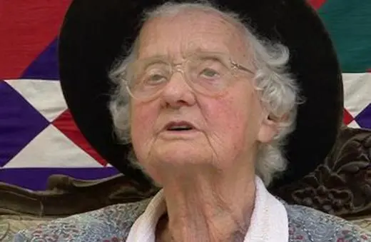 An elderly woman with curly white hair, wearing glasses and a black hat, sits in front of a colorful quilt-like backdrop.