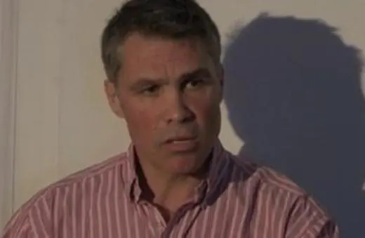 A man with short, grayish hair speaks intently while wearing a red and white striped shirt against a light background.