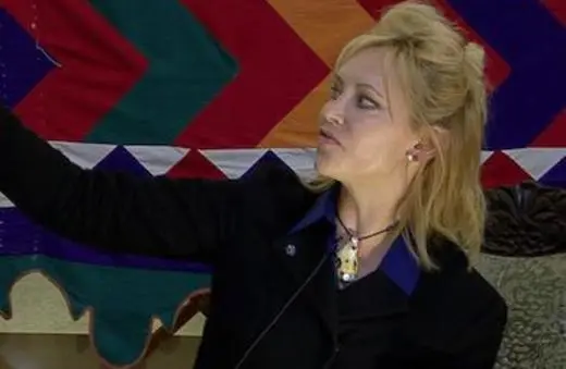 A woman with blonde hair and a black jacket gestures while sitting in front of a colorful patterned backdrop.