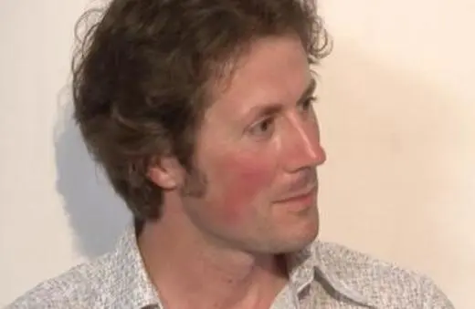 A man with wavy hair, wearing a patterned shirt, looking thoughtfully to his side against a plain background.