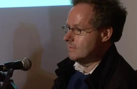 A man with glasses and tousled hair sits at a microphone, appearing contemplative during a presentation or discussion.