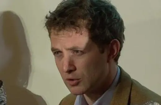A man with curly hair and a serious expression speaks at a microphone during a press conference or public event.