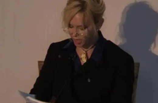 A woman with blonde hair and glasses reading from a paper, seated on a stage with a shadow cast on the wall behind her.