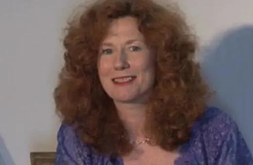 A woman with curly red hair wearing a purple top smiles warmly at the camera.