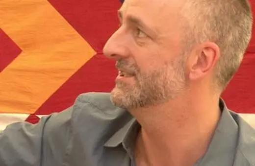 A man with short gray hair and a beard, wearing a blue shirt, smiles while sitting in front of a colorful patterned background.