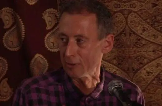 A man speaking at a microphone, wearing a checkered shirt, against a patterned backdrop adorned with paisley designs.