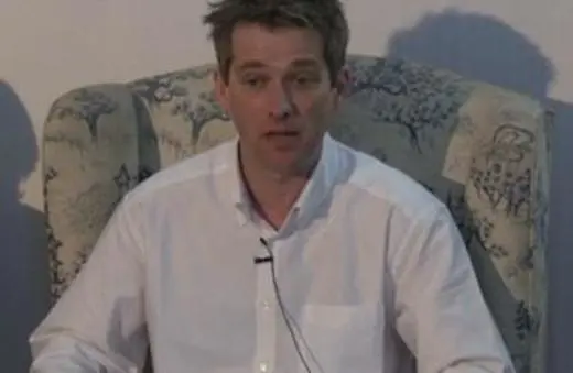 A man in a white shirt sitting on a patterned armchair, engaged in a discussion or lecture.