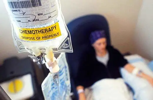 A chemotherapy IV bag hangs in focus, with a patient in a chair blurred in the background, undergoing cancer treatment.