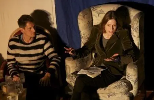 Two individuals engage in a discussion while seated in plush armchairs, with one gesturing expressively and the other attentively listening. Subtle lighting enhances the intimate atmosphere.