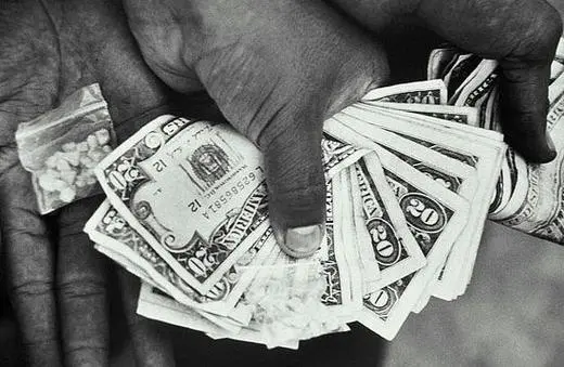 A close-up of hands exchanging cash, including various denominations of bills, alongside a small bag containing a white substance.