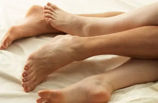 A close-up of two pairs of feet intertwined on a white bedspread, suggesting intimacy and closeness.