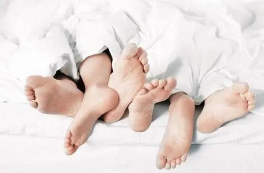 A close-up of several pairs of feet sticking out from underneath a white blanket on a bed.