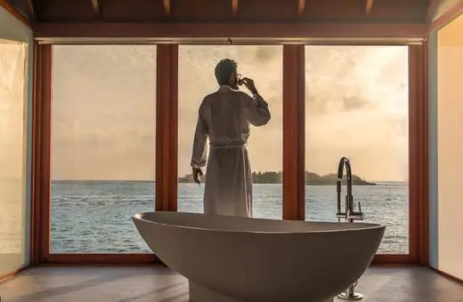 A person in a bathrobe stands by a large window overlooking the ocean, with a freestanding bathtub in the foreground, during sunset.
