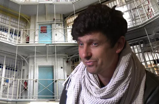 A young man wearing a scarf sits in front of a prison environment, featuring multiple levels and wire mesh elements in the background.