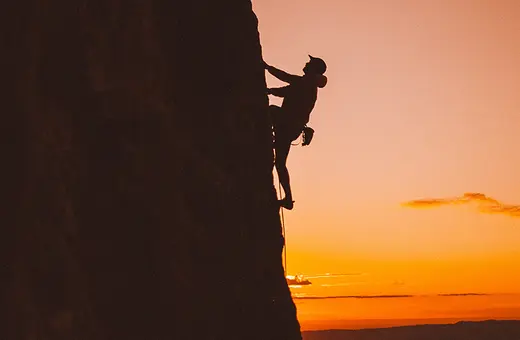 A lone rock climber grips a steep cliff in silhouette against a glowing orange sunset, creating a dramatic, adventurous scene of courage and solitude.