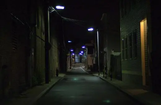 A dark, narrow alley illuminated by street lamps, featuring brick buildings on either side and a faint light at the end of the path.