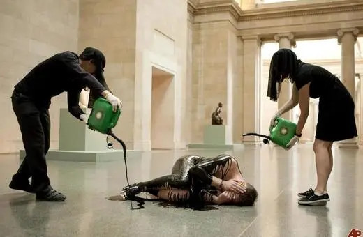 Performance art piece featuring two individuals pouring black liquid over a person lying on the floor in a museum setting, emphasizing themes of vulnerability and transformation.