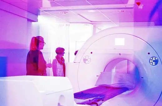 A medical imaging room featuring a CT scanner, with two individuals observing the equipment in a brightly lit environment infused with purple hues.