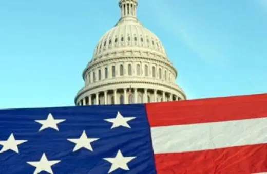 A large American flag draped in front of the United States Capitol building under a clear blue sky.