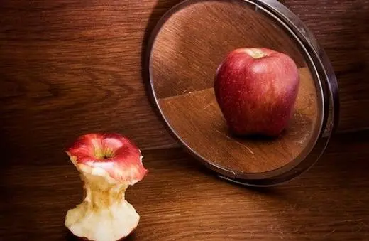 A bite-sized apple core sits next to a whole red apple reflected in a round mirror against a wooden background.